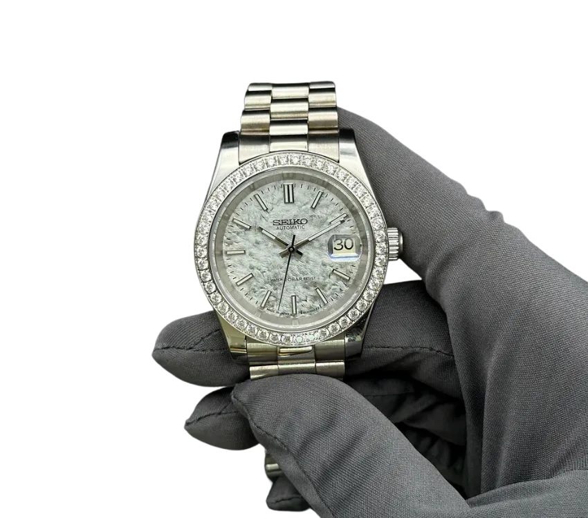 Montre automatique Seiko avec cadran effet pierre texturée, lunette sertie de diamants, affichage de la date, et bracelet en acier inoxydable, tenue par une main gantée grise.