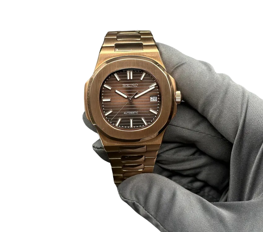 L'image montre une montre automatique Seiko avec un boîtier et un bracelet en métal de couleur bronze. Le cadran affiche un motif horizontal avec des index et des aiguilles en argent, ainsi qu'une fenêtre de date à 3 heures. La montre est tenue par une main gantée pour mettre en valeur sa finition élégante et son design sophistiqué.