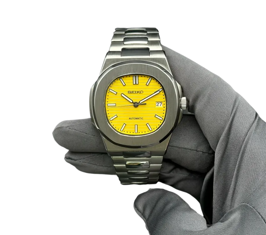 Montre Seiko automatique avec boîtier en acier inoxydable brossé, cadran jaune vif texturé, index et aiguilles luminescents, avec affichage de la date à 3 heures. Présentée dans une main gantée grise.