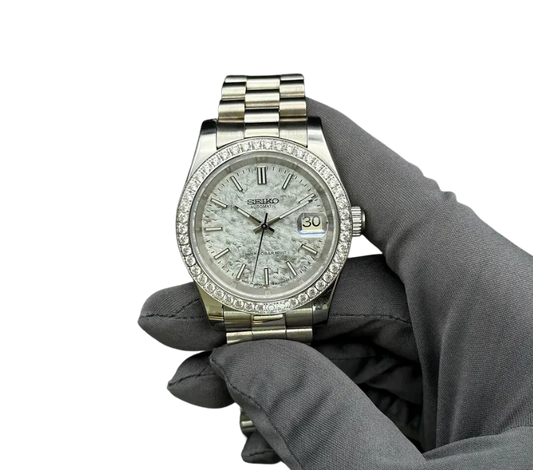 Montre automatique Seiko avec cadran effet pierre texturée, lunette sertie de diamants, affichage de la date, et bracelet en acier inoxydable, tenue par une main gantée grise.