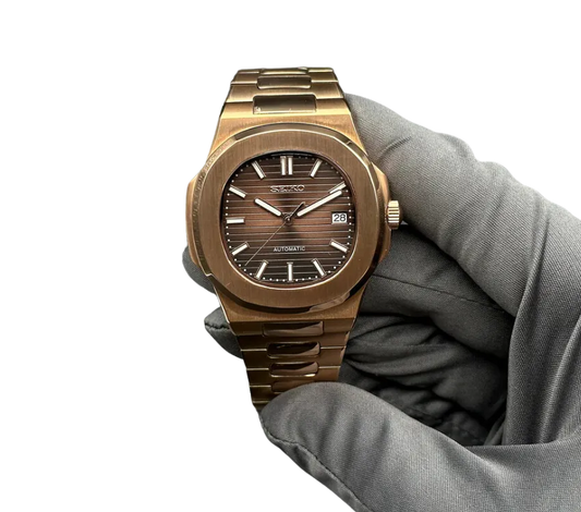 L'image montre une montre automatique Seiko avec un boîtier et un bracelet en métal de couleur bronze. Le cadran affiche un motif horizontal avec des index et des aiguilles en argent, ainsi qu'une fenêtre de date à 3 heures. La montre est tenue par une main gantée pour mettre en valeur sa finition élégante et son design sophistiqué.