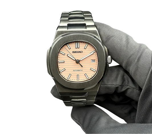 Montre automatique Seiko avec cadran rose clair, boîtier et bracelet en acier inoxydable, tenue par une main gantée.
